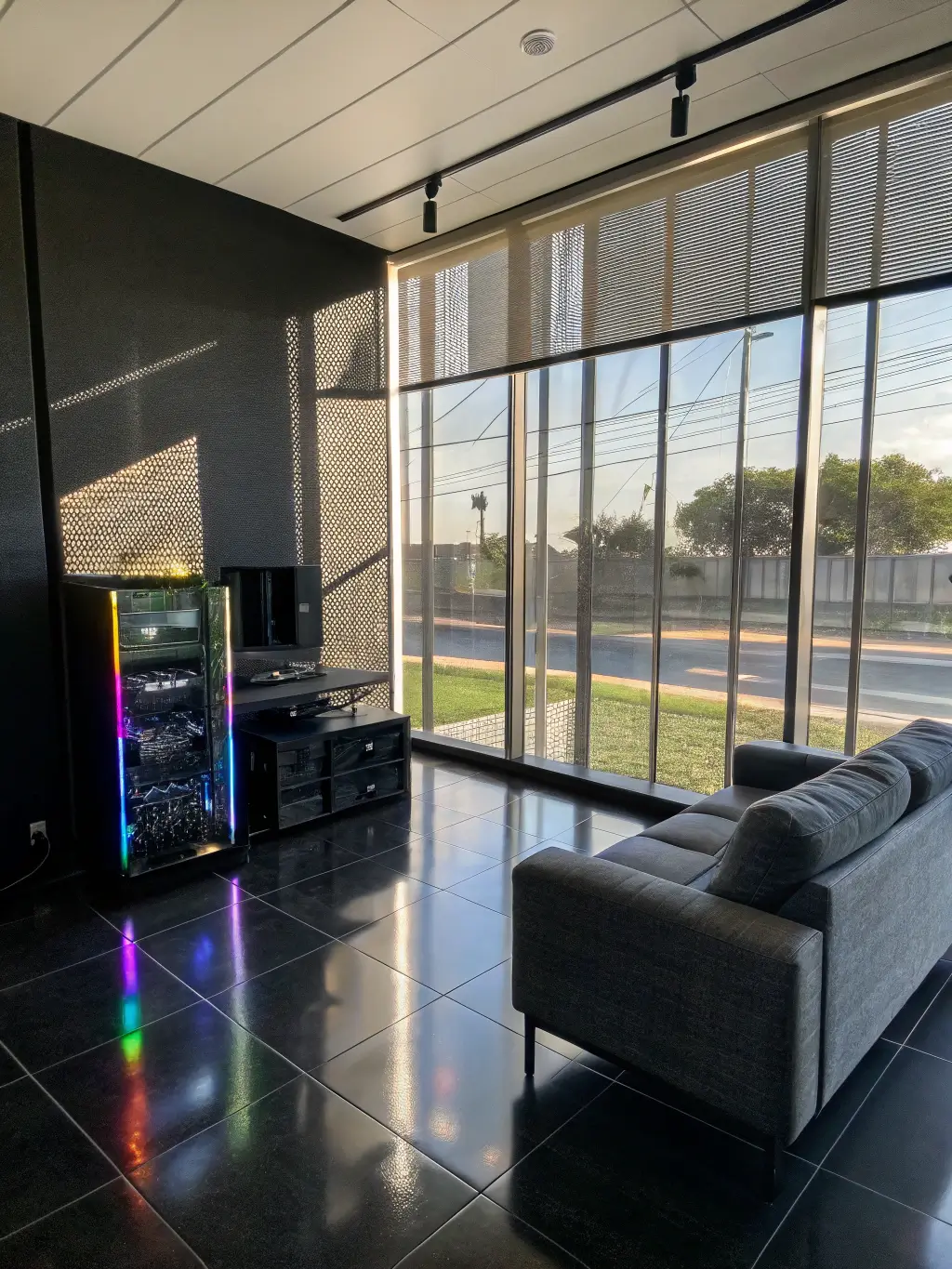 Illuminated gaming lounge with floor-to-ceiling windows, rainbow reflections cast from a transparent PC with custom water cooling, and a central modular sofa on glossy black floor.