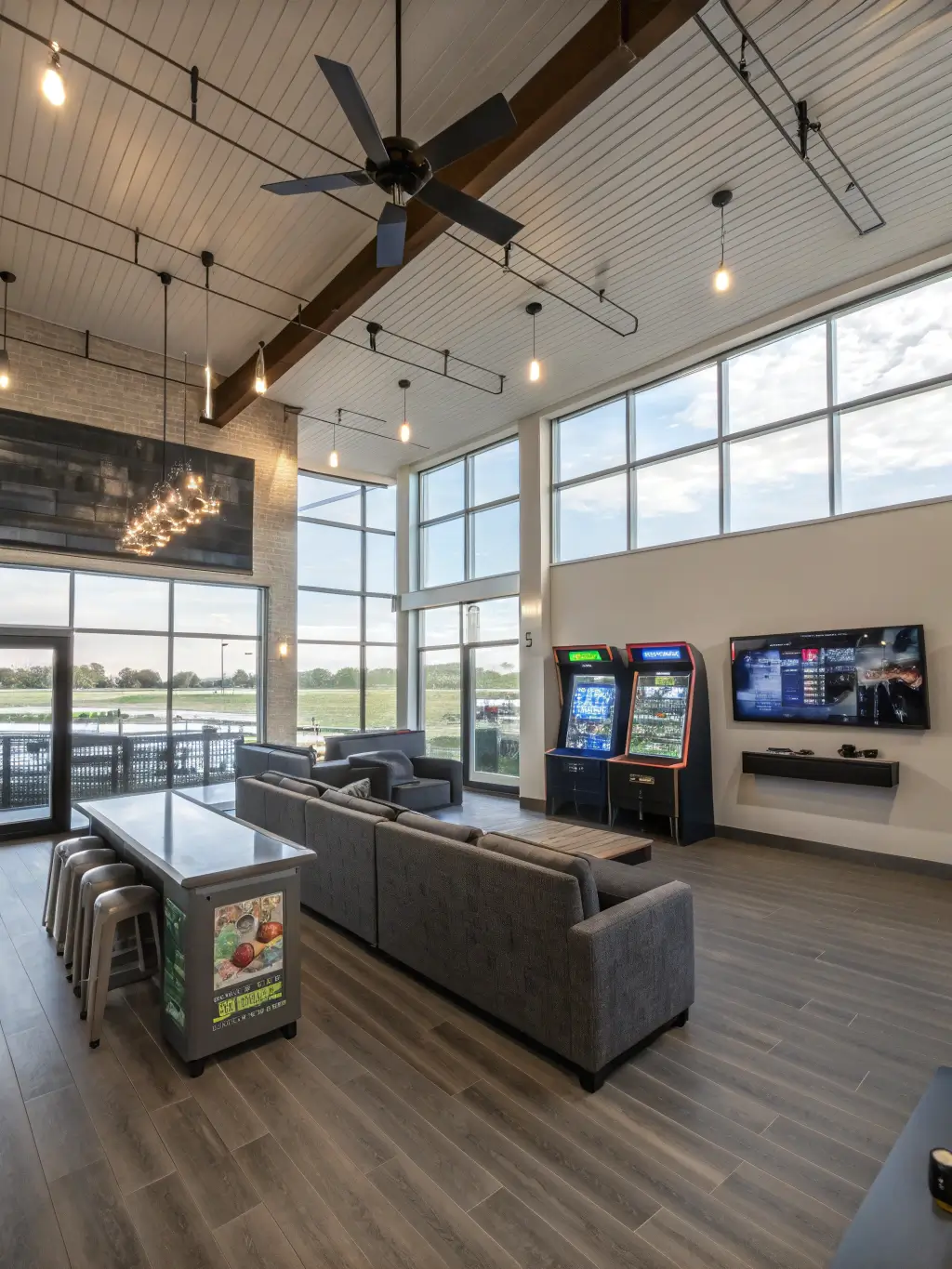 Bird's eye view of a spacious 600sq ft social gaming lounge with panoramic windows, double-height ceiling, modern fan, abstract gaming art on floating panels, and custom console display unit with internal lighting. The area features a conversation pit with modular gaming seating, gray washed engineered hardwood flooring, and pendant cluster lights, showcasing a social layout with a mix of natural and artificial light.